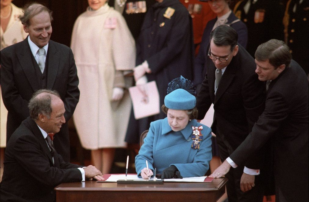 La reine Élisabeth II signe la proclamation de la Loi constitutionnelle, assise à une table avec le premier ministre Pierre Elliott Trudeau. Autour d'eux se tiennent le ministre du Travail Gerald Regan, le greffier du Conseil privé Michael Pitfield et Michael Kirby.